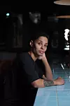 Kvinna med tatuering sitter vid en blå kaklad bardisk i en mörk bar.